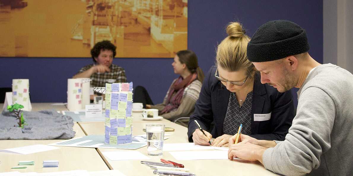 Vier Personen sitzen an einem Tisch. Auf dem Tisch sind kleine Säulen mit Zetteln aufgebaut. Zwei Personen schreiben etwas auf weitere Post Its. Die beiden Personen im Hintergrund unterhalten sich.
