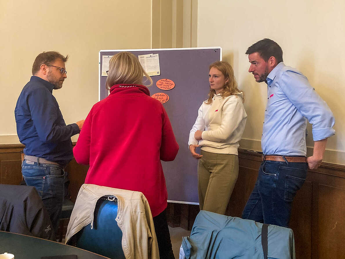 Vier Menschen stehen vor einer Pinnwand und unterhalten sich.