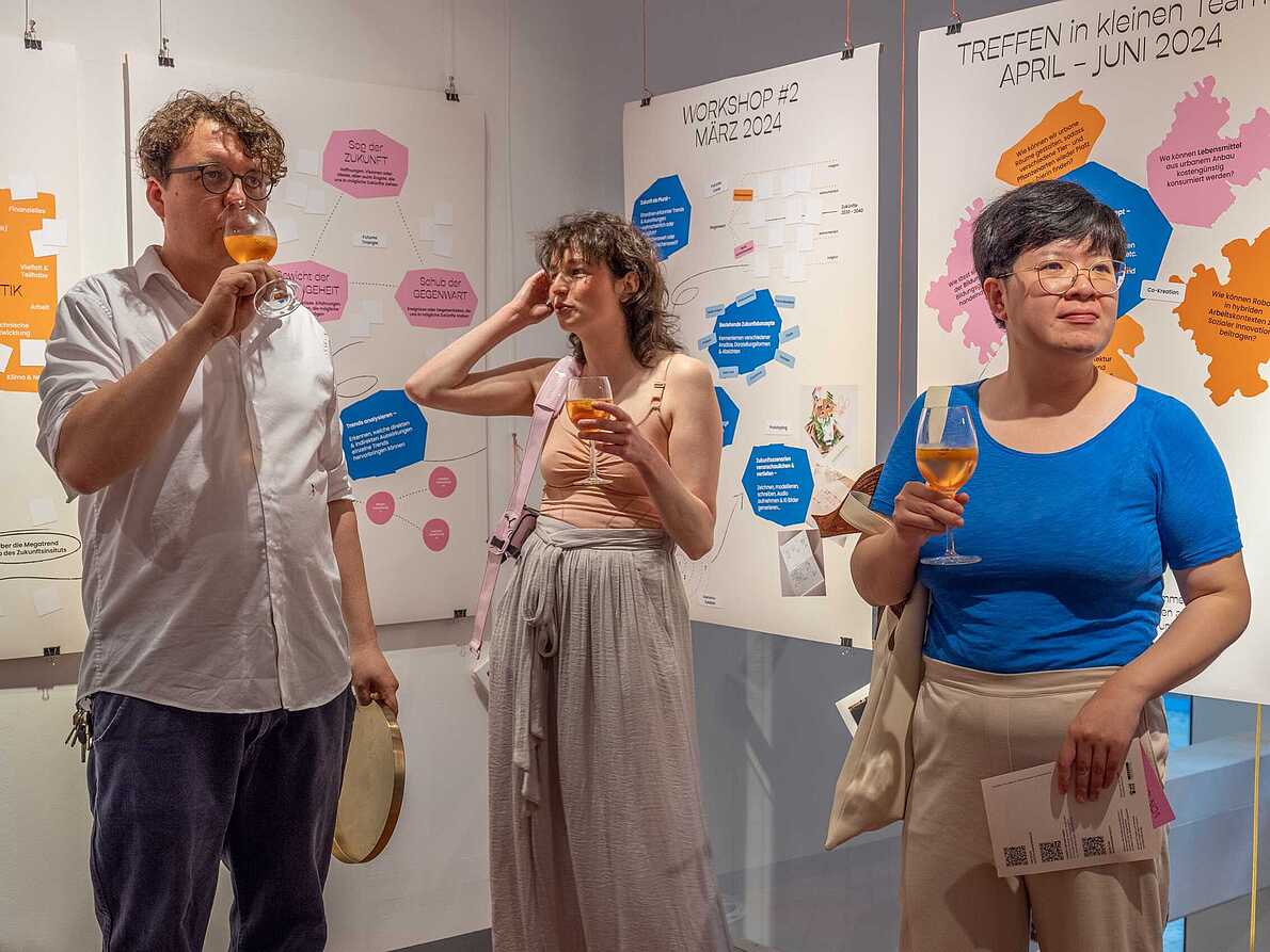 Drei Personen im Gespräch auf der Eröffnung der Ausstellung "Different Futures Make A Dialogue". Hinter ihnen hängen vier große Plakate mit bunten Text-Grafiken