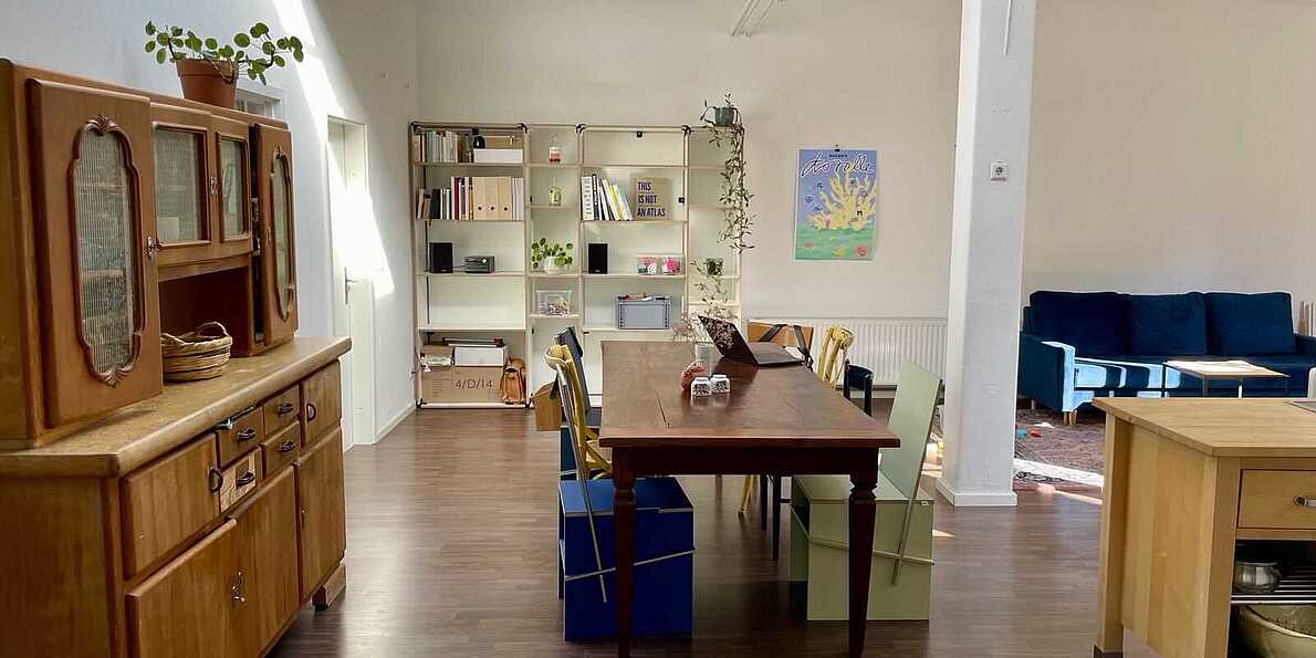 Abbildung des Gemeinschaftsraumes im Büro von koralle: Ein großer, heller Raum, in dessen Mitte, neben einer Säule, ein großer Esstisch mit 10 Stühlen steht.