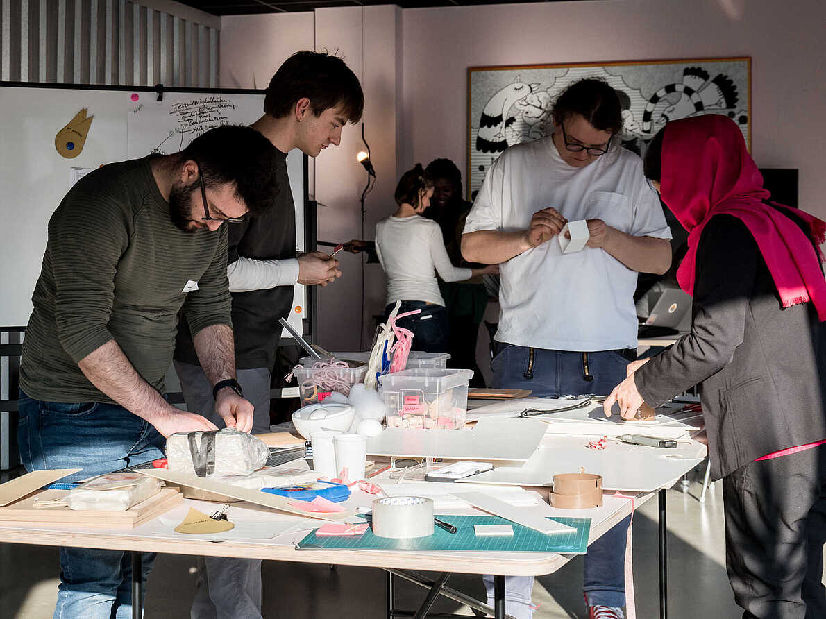 Teilnehmende stehen um einen Tisch und basteln unter anderem mit Ton, Schnur, Holz und Klebeband.