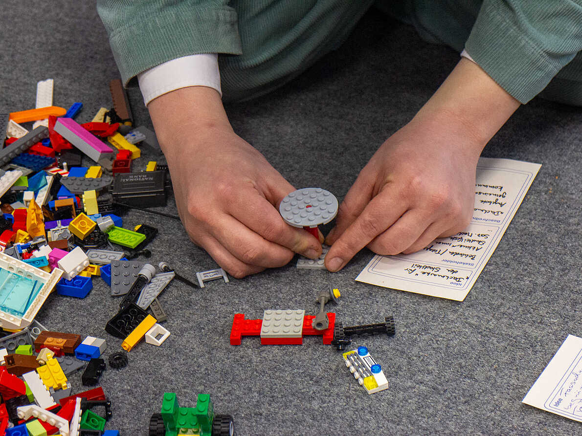 Eine Person sitzt auf dem Boden und baut etwas mit Lego. Detailaufnahme der Hände.