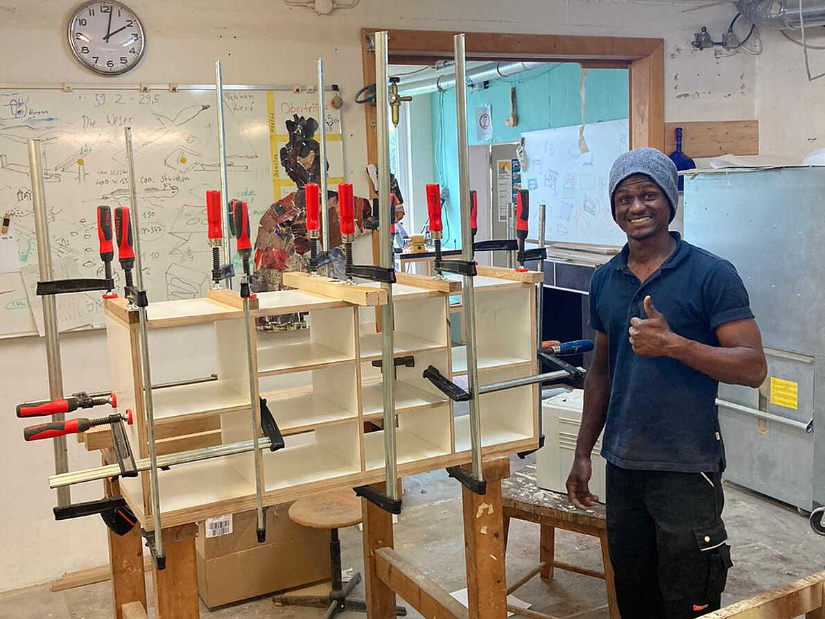 Das Bild zeigt Diallo in der Werkstatt stehend und den Daumen nach oben zeigend. Neben ihm ist der Beginn eines Möbelstücks zu erkennen, das mit vielen Schraubzwingen zusammengehalten wird.