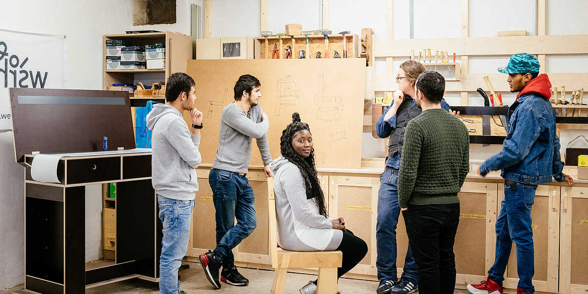 Eine Fotografie von 6 Personen, die auf eine Wand mit Zeichnungen blicken und sich in einem Gespräch befinden. Eine von ihnen sitzt auf einem Stuhl und blickt in die Kamera. Sie befinden sich in der Holzwerkstatt des Projekts weserholz.