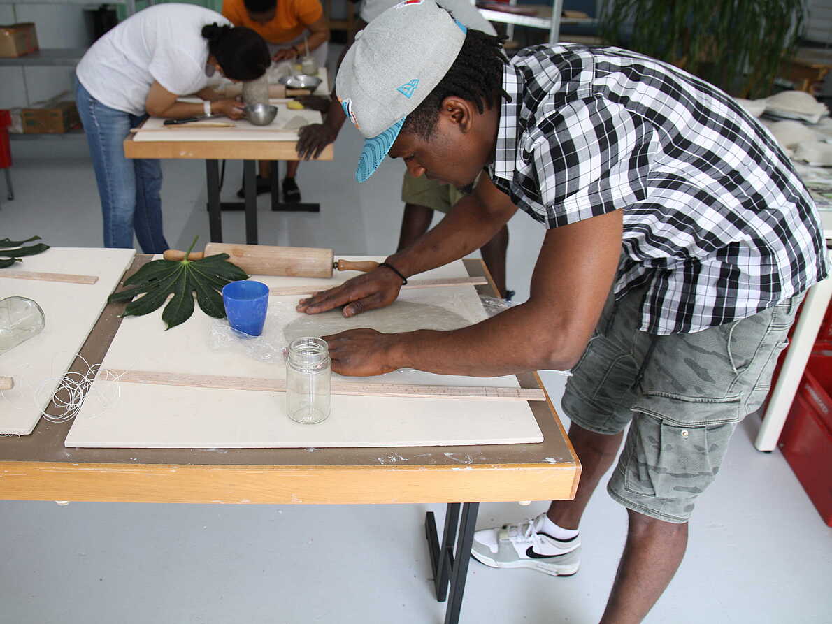 Das Bild zeigt eine Person. Diallo ist vorgebeugt über einen Tisch und arbeitet mit Ton