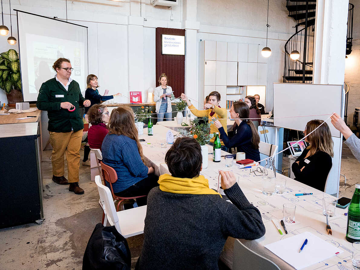 Arbeitssituation während des Workshops "Verwaltung und Soziale Innovation". Workshopteilnehmende sitzen an einem langen Tisch, dessen Tischdecke mit Arbeitsaufträgen bedruckt ist. Die Teilnehmenden halten einen langen Faden in die Luft, welcher sie verbindet.