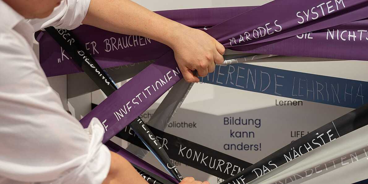 Detail aus dem Szenario "Bildung" der Ausstellung "Different Futures Make A Dialogue": Eine Person zieht breite Gummibänder zur Seite. Auf den Gummibändern stehen verschiedene Wörter. Sie sind auf dem Bild nicht im Detail zu erkennen.
