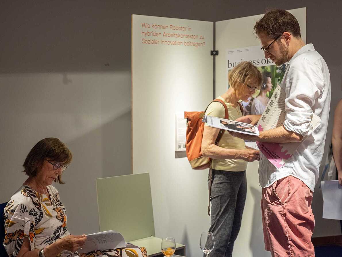 Drei Personen betrachten das Szenario "Arbeit", ein Magazin, auf der Ausstellung "Different Futures Make A Dialogue". Eine von Ihnen sitzt auf einem Stuhl und liest in dem Magazin. Ihr gegenüber steht eine Person und liest in dem Magazin. Hinten steht eine weitere Person vor der Stellwand mit Plakat und liest den Infozettel des Szenarios.