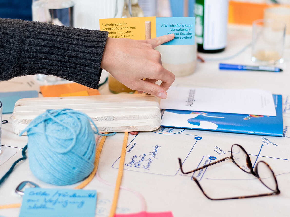 Arbeitssituation beim Workshop "Verwaltung und Soziale Innovation". Zu sehen ist eine Hand, welche auf einen Zettel zeigt. Auf dem Zettel steht die Frage "Welche Rolle kann Verwaltung im Kontext Sozialer Innovation spielen?"