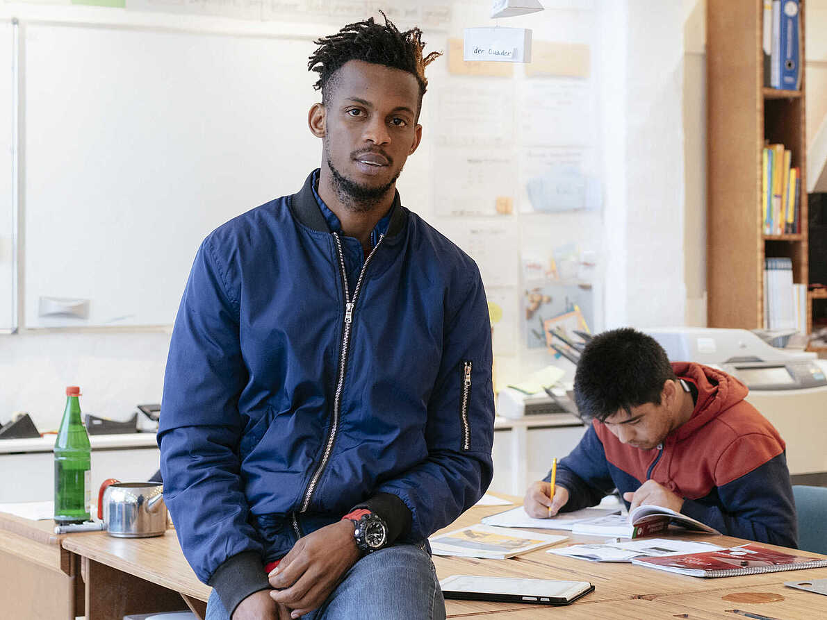 Eine Person sitzt lehnend an einem Tisch und schaut in die Kamera. Eine andere Person im Hintergrund schreibt etwas in ein Heft.