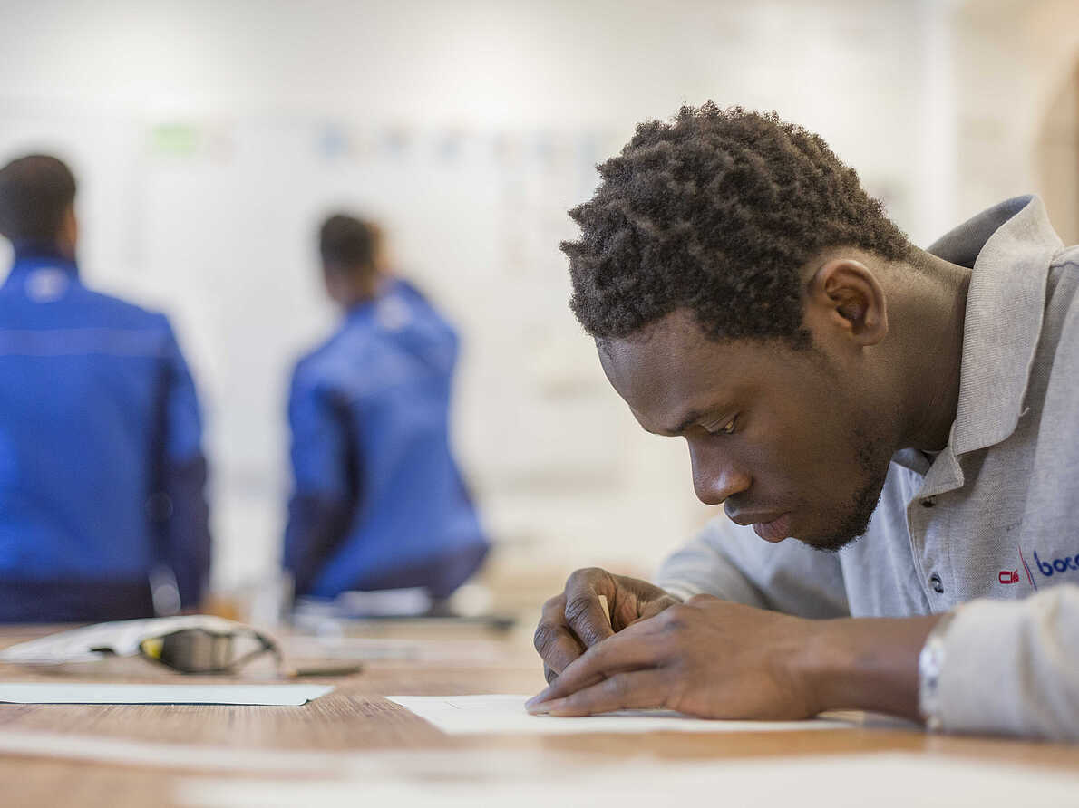 Eine Person sitzt am Tisch und zeichnet. Sie sieht dabei sehr konzentriert aus. Im Hintergrund stehen zwei Personen in Arbeitskleidung an einem Whiteboard