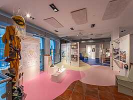 Blick in den Ausstellungsraum von "Different Futures Make A Dialogue". Links hängt ein gelber Overall mit Helm und Netz von der Decke. Auf dem Fußboden kleben verschiedenfarbige Flächen. Es sind weiße Stellwände aufgestellt, die Texte und verschiede, im Detail nich erkennbare, Dingen enthalten.