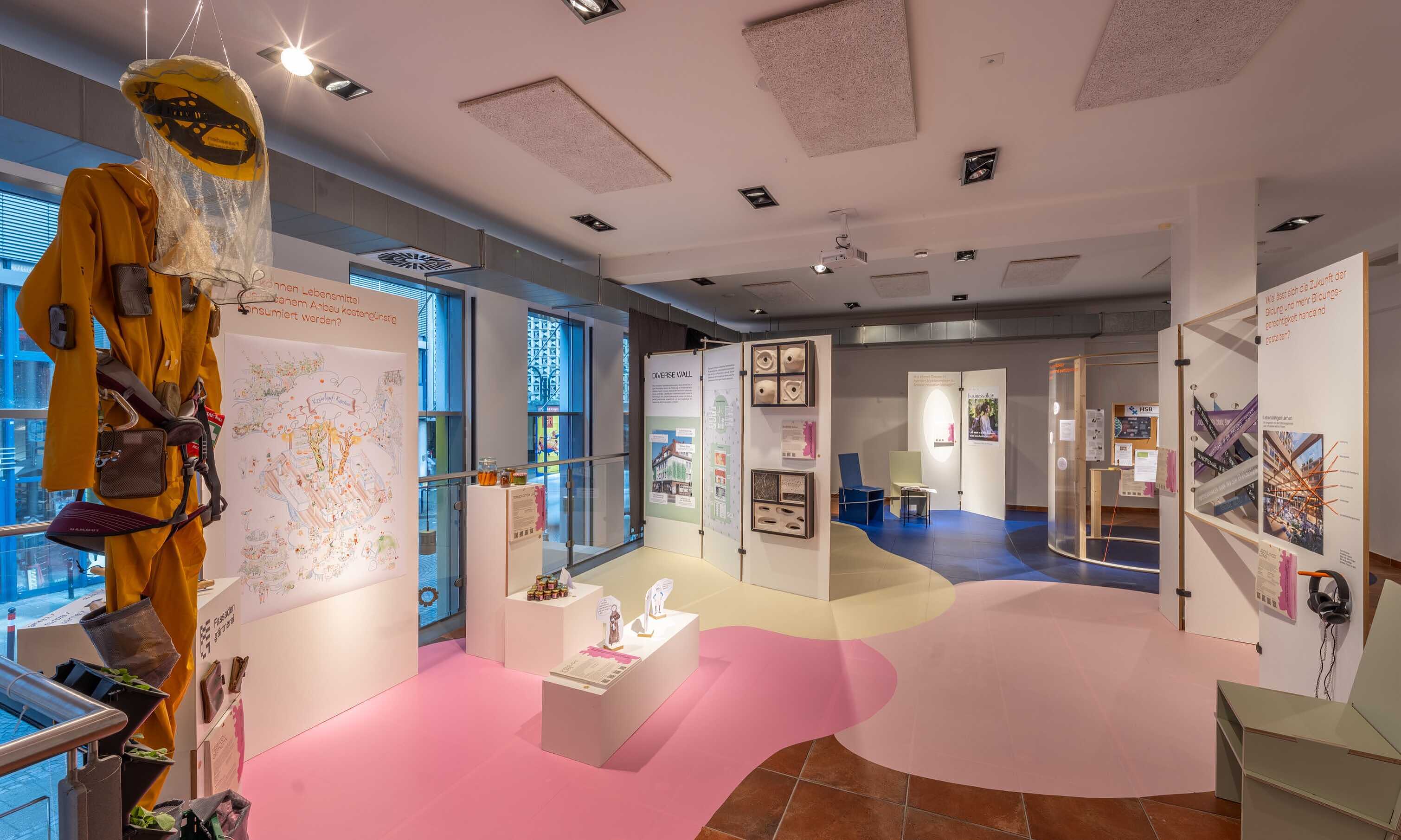 Blick in den Ausstellungsraum von "Different Futures Make A Dialogue". Links hängt ein gelber Overall mit Helm und Netz von der Decke. Auf dem Fußboden kleben verschiedenfarbige Flächen. Es sind weiße Stellwände aufgestellt, die Texte und verschiede, im Detail nich erkennbare, Dingen enthalten.