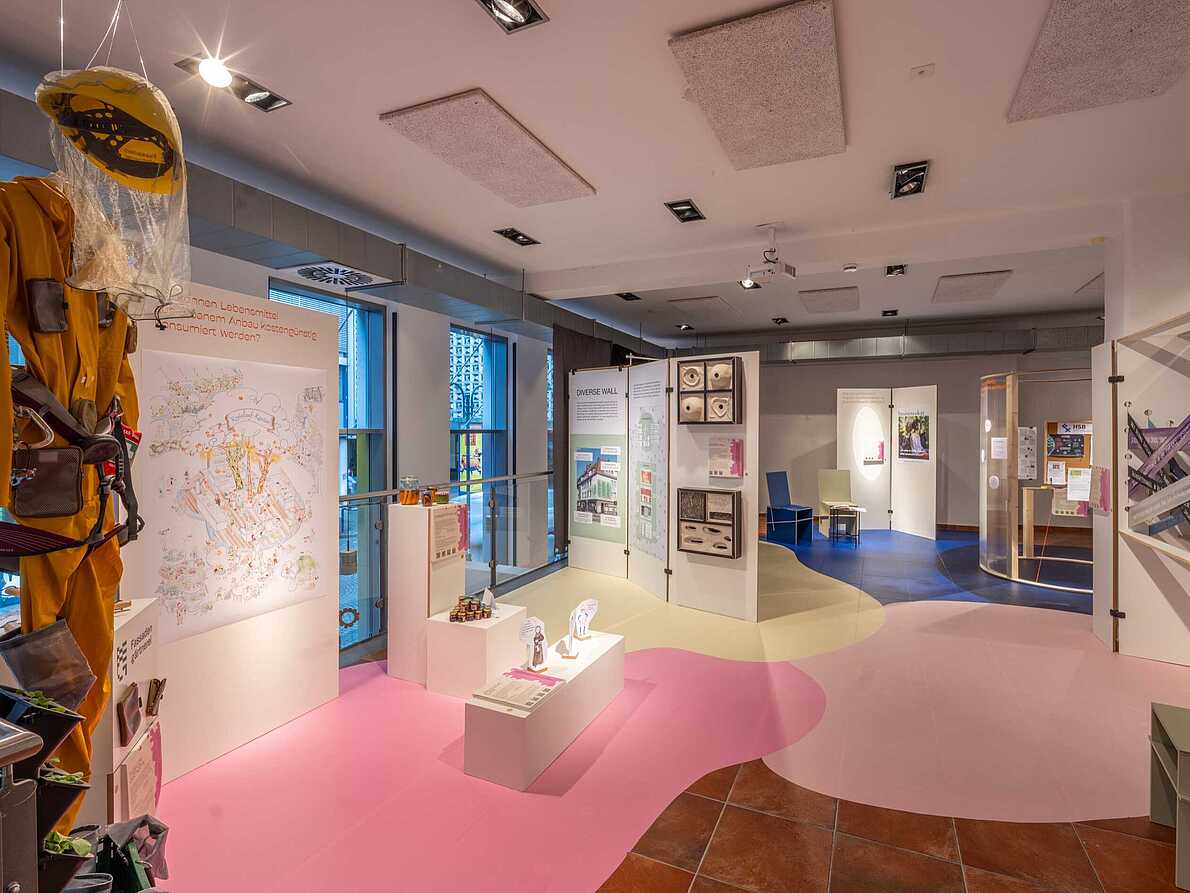 Blick in den Ausstellungsraum von "Different Futures Make A Dialogue". Links hängt ein gelber Overall mit Helm und Netz von der Decke. Auf dem Fußboden kleben verschiedenfarbige Flächen. Es sind weiße Stellwände aufgestellt, die Texte und verschiede, im Detail nich erkennbare, Dingen enthalten.